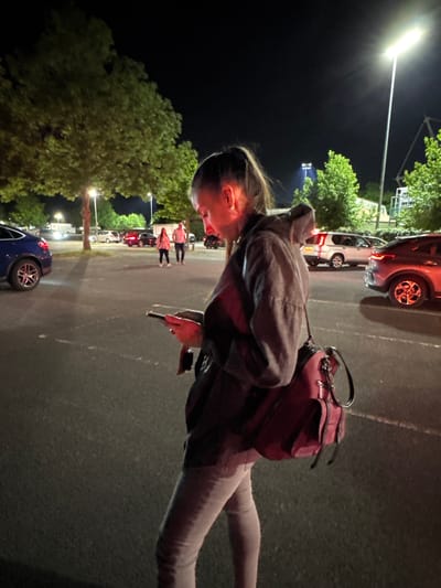 The image shows a woman standing in a parking lot at night. She is wearing a hooded jacket, jeans, and a backpack. She is looking down at a phone in her hand. The parking lot is asphalt and has several cars parked in it. Trees are visible on the edge of the parking lot. There are other people visible in the distance. It is not possible to confirm the exact location as the Borough of Spelthorne, United Kingdom without further information or landmarks in the image.