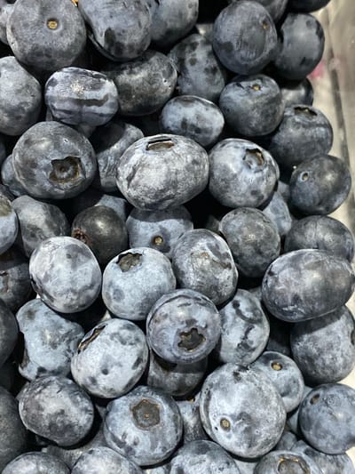 The image displays a close-up, overhead view of a pile of blueberries. The berries are predominantly a dark blue to purplish hue with some appearing lighter due to a powdery bloom on their surface. They are irregularly shaped, mostly spherical or slightly oval, and are clustered together. Some berries show the characteristic small indentation at the top where the stem was attached, often revealing a lighter brown or yellow center. The lighting suggests an indoor setting, likely a kitchen or grocery store display, with no visible background elements that would indicate a specific location. The image does not provide any information about the city of Kushchovskaya, Russia, nor does it contain any people or identifiable actions.