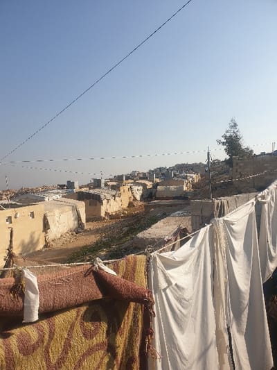 This media file captures an outdoor scene under a clear, bright blue sky, likely in the area of Killi, Syria. The image is oriented on its side but depicts a horizontal landscape.
In the foreground, two large white canvas tarpaulins or tent-like structures are prominently visible on the right side, appearing to be part of a temporary shelter or covering. Below them, two rugs or blankets are laid out or rolled. One is a solid reddish-brown, and the other features a distinctive yellow and brown patterned design, possibly drying in the sun or stored outdoors. Ropes and ties are visible, securing these items.
The middle and background reveal a sprawling settlement of numerous low-rise buildings, predominantly constructed from light-colored materials, suggesting a mix of concrete, stone, or mud brick. These structures appear simple and somewhat weathered, indicative of basic living conditions. A narrow, dark path or channel, possibly a dry watercourse or track, runs through the settlement. A lone tree with green foliage can be seen on the distant horizon, adding a touch of natural life to the otherwise arid-looking landscape. Overhead, a long, thin dark line, likely a utility wire or cable, stretches across the expansive sky.
The scene appears to take place during broad daylight, possibly midday or early afternoon, given the harsh shadows and the intense brightness of the sky, indicating sunny and dry weather conditions. No people are visible in the image. The presence of makeshift shelters and the general appearance of the settlement align with the context of a residential area or camp in a region like Killi, Syria, often impacted by displacement or conflict. There is no visible text in the image.