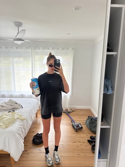 A mirrored view depicts a woman in a black tee and shorts, holding a carton of coconut milk. She wears sunglasses and trainers with black socks. The bedroom has wooden floors, a white bed with clothes strewn on it, and a Dyson vacuum cleaner. Overhead is a wardrobe with items stored on the shelves. A ceiling fan is visible. The reflection conveys a casual, everyday scene.