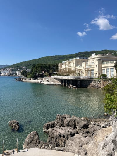 The image presents a serene coastal vista of Grad Opatija, Croatia, under a brilliant blue sky dotted with scattered white clouds. The scene is bathed in bright daylight, suggesting late morning or early afternoon.
The majority of the lower and right frame is occupied by the clear, calm waters of the Adriatic Sea, which exhibits a beautiful turquoise-green hue. Sunlight reflects off the water's surface, creating intricate rippling patterns. Along the immediate shoreline, large, dark grey rocks are visible, both submerged in the clear water and exposed on the bank. Sections of the coast feature built-up stone retaining walls and concrete edges, from which metal ladders extend into the sea, indicating a popular swimming spot.
On the left side of the frame, a lush, densely forested hillside rises steeply, forming a verdant backdrop to the town. Prominently featured in the mid-ground is a grand, multi-story building with a light-colored, possibly cream or pale yellow, facade. This elegant structure, identified by the visible text "GRAND HOTEL WAGNER" on its front, showcases classical architectural details such as numerous windows, balconies with white railings, and decorative elements including statues along its roofline. Further along the coastline to the left, additional buildings are visible nestled amidst the greenery, suggesting a vibrant resort town built into the hillside.
The sky is a vivid, cloudless blue for the most part, though some fluffy white clouds are present in the upper left, and a faint airplane contrail streaks across the upper right. The overall atmosphere conveyed is one of tranquility, natural beauty, and a pleasant holiday setting, devoid of explicit human activity but implying leisure and relaxation.