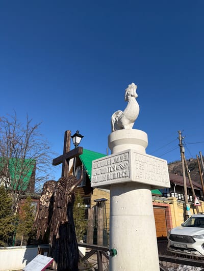 Here is a description of the image:
The image shows a monument in the city of Manzherok, Russia, featuring a white statue of a rooster atop a stone pillar inscribed with text in Russian. Below the rooster monument is a wooden sculpture of an angel attached to a cross with a lamppost. A sign with more Russian text and the price "4000 rub" sits next to the angel. The background includes buildings, power lines, trees, a blue sky, and a portion of a white car with the license plate "K164EY 122".
