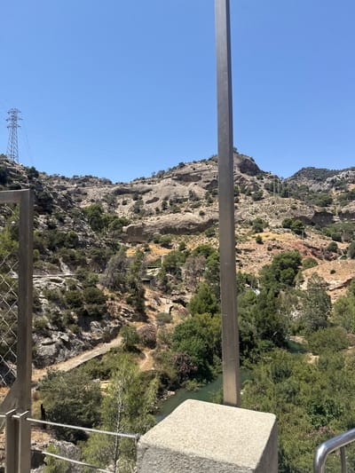 The image depicts a scenic view of a gorge in Ardales, Spain, likely at a high vantage point. The dominant subjects are the steep, rocky cliffs covered in sparse vegetation, with a winding river flowing through the gorge below. Lush green trees and shrubs cling to the mountainside, creating a vibrant contrast with the arid rock faces.

In the distance, a narrow pathway is visible, carved into the side of the gorge, suggesting a hiking trail or a part of the famous Caminito del Rey. A few small structures or ruins are nestled into the cliffs. There are hints of human activity, with what appear to be a few people visible on the pathway below, and a visible power line structure in the background, suggesting human infrastructure in the natural landscape.

The time of day appears to be mid-morning or early afternoon, indicated by the bright, clear blue sky and the strong sunlight casting distinct shadows on the rocky terrain. The weather is sunny and fair, contributing to the picturesque and inviting atmosphere of the location. The overall mood is one of natural beauty and rugged grandeur, with a touch of human presence harmonizing with the environment. The perspective suggests the photo was taken from a bridge or walkway overlooking the gorge.