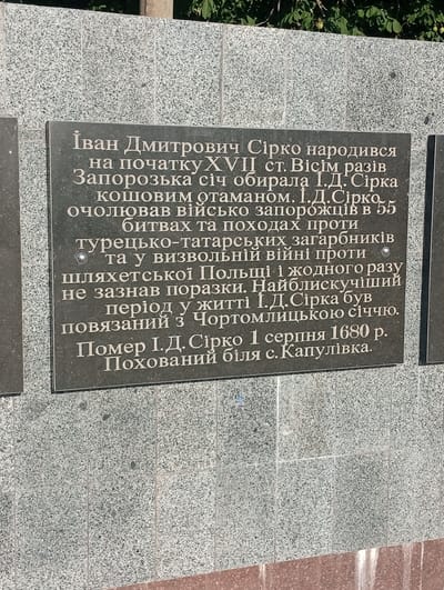 This is a photograph of a memorial plaque mounted on a granite wall. The plaque is dark grey with carved text in a lighter color. The text appears to be in Ukrainian and describes Ivan Dmitrovich Sirko, a military leader. It states that he was born at the beginning of the 17th century, was elected Koshovyi Otaman of the Zaporizhian Sich eight times, and led Zaporozhian Cossacks in 55 battles and campaigns against Turkish-Tatar aggressors and in the liberation war against noble Poland, never suffering defeat. The plaque mentions that the most brilliant period of his life was connected with the Chortomlyk Sich and that he died on August 1, 1680, and was buried near the village of Kapulivka.
The background of the plaque is a rough-textured granite wall, possibly part of a larger monument or building. Above the plaque, the top of the granite wall is visible, with some green foliage from trees suggesting an outdoor setting, likely during daylight hours with fair weather. The image is taken at a slightly downward angle, focusing on the memorial. The inscription is the primary subject of the photograph, serving to commemorate Ivan Sirko and his historical significance. The overall tone is serious and historical.