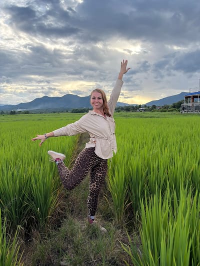 In Ban Pong, Thailand, a woman stands in a lush green rice field under a cloudy sky. She is posing with her left leg bent at the knee and extended backward, while her right arm is raised and her left arm is extended to the side. The woman is wearing a tan collared shirt with cinched waist and leopard-print leggings. The background shows a mountain range and a building with a blue roof. The rice paddy is dense with vibrant green stalks, and a narrow dirt path is visible where the woman is standing.