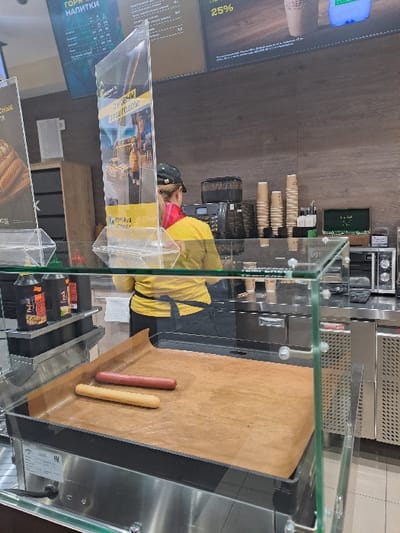 A glass display case contains two hot dogs on a brown paper-lined tray. One hot dog is pinkish-red, the other is a pale yellow color. Behind the case, a person in a yellow and black uniform stands near a series of paper cups. An advertisement is attached to the front of the display. A menu board is visible in the background. Appliances appear behind the cups.