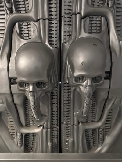 The image shows a close-up of two sections of a silver-colored, bio-mechanical relief. The relief features skull-like forms with elongated features and ribbed/toothed sections. The piece's style is reminiscent of H.R. Giger's biomechanical artwork. These reliefs are part of the Giger Bar in Epagny, Switzerland.
