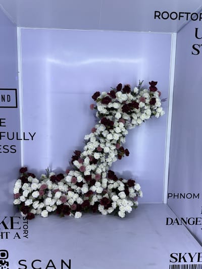 This media file captures a corner of an indoor space, likely a venue in Khan Boeng Keng Kang, Cambodia, adorned with a large floral arrangement. The arrangement is shaped like a number "1" and is composed of numerous white and dark red roses, interspersed with smaller pinkish-purple floral accents and sprigs of lavender. The flowers are densely packed, creating a rich and textured display against a pale purple backdrop.
The background wall is a smooth, light purple surface, reflecting light and giving the impression of a studio or event space. Towards the left and top, black text is visible on a lighter surface, oriented vertically. This text reads "ROOFTOP", "U", and "S". Towards the right and top, more vertical text is present, reading "PHNOM", "DANGE", and "SKYE" with a barcode below "SKYE". At the bottom, horizontal text reads "STORY", "KEY", "HT A", and "ND", along with an icon that appears to be a QR code.
The scene appears to be set up for an event, possibly a celebration or a photo opportunity, given the decorative floral installation and the presence of branding or directional text. The lighting is even and soft, suggesting an indoor setting without direct sunlight. There are no people visible in the image, focusing the viewer's attention entirely on the floral art and the textual elements. The overall mood is one of understated elegance and visual interest, driven by the contrast between the organic beauty of the flowers and the graphic nature of the text.