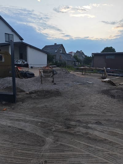 The image depicts an outdoor construction site in Vasilkovo, Russia, under a cloudy sky. In the foreground, there is a patch of sand with tire tracks. A pile of grey gravel is to the left, and a concrete mixer stands next to a pile of sand. Behind these, a white modern house with dark trim and a dark roof is visible. Further back, several older, grey houses with pitched roofs are scattered among trees. A dark brown building with a barred window is also present on the right side of the image. Wooden planks and other construction materials are visible throughout the scene. No people are present.
