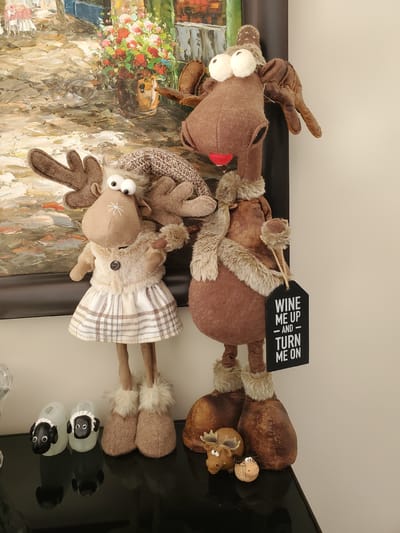 The media file displays two stuffed reindeer figures, likely Christmas decorations, placed on a reflective surface against a light-colored wall. The reindeer are brown and appear to be made of fabric and faux fur. The larger reindeer on the left has a red nose and two large white pom-pom eyes. It is wearing a brown knitted hat with antlers. A black tag hangs from its neck with white text that reads "WINE ME UP - AND TURN ME ON".
The second, smaller reindeer to the right is wearing a plaid dress and also has large white pom-pom eyes and antlers. Its legs are made of brown fabric, and its feet are covered in fluffy material. Beside the reindeer are two small ceramic sheep with black faces and ears. The background behind the reindeer features a colorful painting or tapestry with floral elements and abstract brushstrokes, possibly depicting a street scene. The overall impression is a festive, indoor display, likely in a home, given the presence of decorative items. The lighting suggests an indoor setting during the daytime. There are no explicit weather or time-of-day indicators, but the overall mood is whimsical and playful. The location context of Kitchener, Canada, provides a geographical setting for where this image might have been taken.