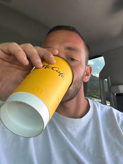 A male subject is depicted inside a vehicle, in what is identified as Agoura Hills, United States of America. He is seen consuming from a yellow McCafé branded paper cup, holding it to his mouth with his right hand. The cup is tilted upwards, suggesting it is being emptied, and the lid is absent. The subject has short, dark hair and a beard, wearing a white crew-neck t-shirt. A distinct tan line or sunburn is visible across his forehead. Through the vehicle's passenger side window, a mountainous terrain with green foliage under a blue sky with white clouds is visible in the background. The car interior features a light gray headliner and door panel.