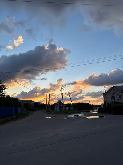 The image depicts a quiet, rural street in Rossosh, Russia, during a sunset. The sky is filled with dramatic clouds, some dark gray and others illuminated with vibrant orange and yellow hues from the setting sun, creating a striking contrast against the clear blue sky. Electrical wires crisscross the sky, supported by utility poles that dot the scene.
On the left side of the street, a blue fence encloses a property with trees. Further down, houses are visible nestled among vegetation. In the center of the road, several puddles of water reflect the colorful sky, indicating recent rainfall.
On the right side of the street, a two-story brick house with white window frames stands prominently. More houses and trees line the street in the distance, leading towards the horizon where the sunset is most intense. The road itself appears to be asphalt, with some cracks and unevenness. The overall atmosphere is peaceful and serene, capturing a moment of natural beauty in a rural setting. There are no people or animals visible in the image.