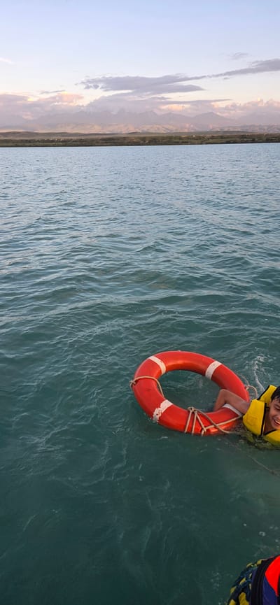 The image depicts a young boy wearing a yellow life jacket, smiling as he holds onto a bright red life preserver in the water. The boy's legs are visible as he floats. The water is a deep teal color with visible ripples and waves, suggesting it is a natural body of water. In the background, a coastline with mountains and a soft, cloudy sky can be seen. The sky suggests it might be sunset or sunrise due to the muted colors and soft light. The overall scene conveys a sense of joy and perhaps a moment of recreation or a lesson in water safety. There is no visible text in the image.