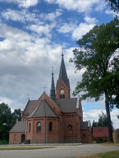The image depicts a brick church with tall spires and crosses against a cloudy sky. A large tree with green foliage is visible in the foreground to the right of the church. The church has arched windows and a clock on one of its spires. A fence separates the church grounds from a paved area in front of it. The scene appears to be an exterior view of a religious building in a town.