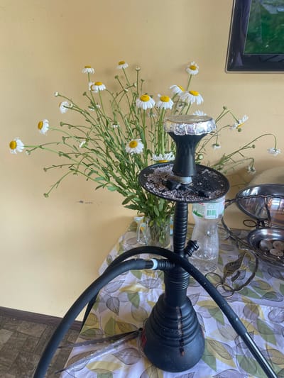 The image depicts an indoor scene in Petrivka, Ukraine. A black hookah is centrally positioned on a table, with its bowl covered in perforated aluminum foil and its charcoal tray containing ash and several black charcoal pieces. Adjacent to the hookah is a clear plastic water bottle. The table is covered with a white tablecloth adorned with a pattern of green and yellow leaves. To the right of the hookah, a decorative metal stand with a leaf design holds small metal bowls, one of which contains several light-colored, irregularly shaped objects. Behind the hookah and bottle, a clear glass pitcher serves as a vase for a substantial bouquet of white daisies with yellow centers and long green stems. The background consists of a textured, light yellow-ochre wall, displaying minor scuffs. In the upper right corner, a portion of a dark-framed television screen is visible, reflecting a green outdoor scene. The floor in the lower-left portion of the frame is tiled with dark, square-patterned tiles. No individuals are present in the image.
