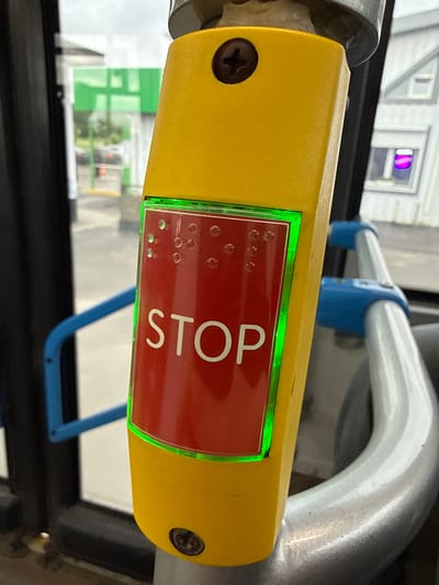 The image shows a close-up of a yellow stop request button on a public transport vehicle, likely a bus, in Yakovlevskoye, Russia. The button is rectangular with a red insert that has the word "STOP" written in white letters. Above the word "STOP" are Braille dots. A bright green LED light outlines the red insert, indicating the button is active. The button is attached to a metallic pole, and a blue grab handle is visible in the background. The environment outside appears to be a street scene with buildings and vehicles visible through the windows. The overall lighting suggests an overcast day. There are no people visible in the image.