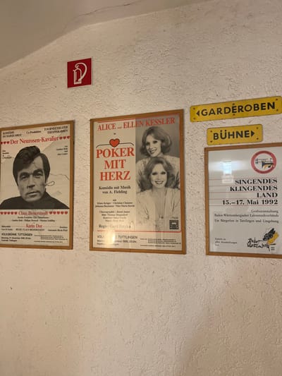 A wall exhibits theatrical posters. One features "Alice and Ellen Kessler in Poker mit Herz" (Poker with Heart). Another promotes "Der Neurosen-Kavalier" (The Neurotic Cavalier) starring Claus Biedderstaedt and Karin Dor. An emergency exit sign is also present. Two yellow directional signs, one for "Garderoben" (wardrobe) and the other for "Bühne" (stage), appear. The final poster advertises "Singendes Klingendes Land" (Singing Sounding Land) music festival, dated May 15-17, 1992, organized by the Baden-Württembergischer Laienmusikverbände (Baden-Württemberg Amateur Music Associations).