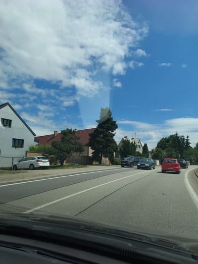 This is a vertical, eye-level, medium shot captured from a vehicle's interior, looking forward on a sunny day with scattered clouds. The scene depicts a residential street in Dražice, Czechia. On the left, a two-story white house with dark windows and a grey roof is visible behind a metal fence. Next to it, a white station wagon is parked on the side of the road. Further down the street, two dark-colored sedans are traveling in the same direction as the camera. Ahead, a red hatchback is also in motion, followed by another dark vehicle. The road is asphalt with white lane markings. Trees and residential buildings line the right side of the road.