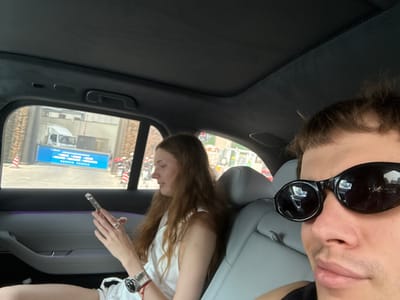 The image depicts a selfie taken inside a car, with two individuals visible. The person in the foreground wears sunglasses and is positioned in the left side of the frame. Behind, another person is seated in a light-colored seat, wearing a white shirt. This person is holding a thin, clear object in their hand, and their hair is brown. Through the car window, a street scene is visible, showing buildings, vehicles, and signage. The sign visible through the car window is in front of a white truck. Based on the Chinese characters on the sign visible in the reflection, the location is identifiable as Hongkou District, Shanghai, China. The car interior is dark, with a visible sunroof.