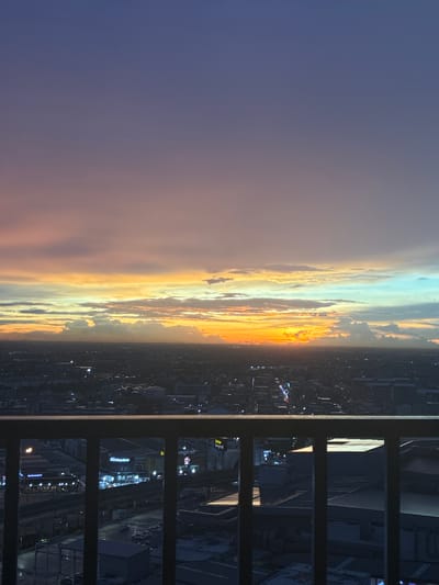 An elevated, wide shot captures a city skyline at dusk. The sky displays a gradient of colors, transitioning from a deep purple at the top to hues of pink, orange, and yellow near the horizon where the sun is setting. Below the colorful sky, a dense urban landscape stretches out, dotted with buildings and streetlights. In the foreground, a dark metal railing divides the view of the city. The city lights are visible in the distance, suggesting a bustling urban environment. The crops show a close-up of the sky, a wider view of the sunset over the city, and a view of the city through the railing, highlighting specific buildings like McDonald's. Based on the provided information and the visual elements, the scene depicts the city of Sao Thong Hin Town Municipality, Thailand, during sunset.