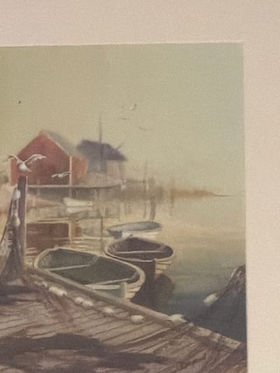 This is a photograph of a watercolor painting depicting a scene at a dock with several boats. In the foreground, a wooden dock is visible, leading to two boats in the water. One boat is mostly in shadow, while the other is lighter and appears to be empty. Further back, more boats are present, and behind them, a red building with a dark roof is situated on stilts over the water. Two birds are in flight near the building, one of which is perched on a wooden post in the left foreground. The background is a hazy representation of the sky and water, with subtle indications of other structures. There is no indication of the city of Villanova d'Albenga, Italy in this image.