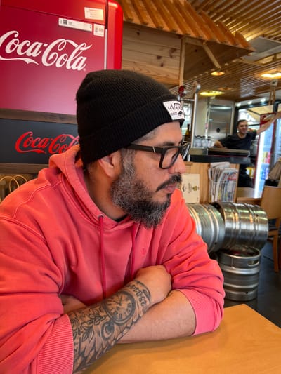 The image shows an indoor scene in what appears to be a cafe or restaurant. A man with a beard, wearing a black beanie and glasses, is resting on a wooden table. He is wearing a pink hoodie and has a clock tattoo on his left forearm. Behind him, there is a red Coca-Cola refrigerator and stacks of beer kegs. In the background, a man wearing black shirt stands behind a counter. The cafe is located in Granollers, Spain.