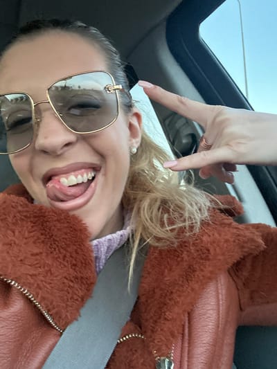 A woman wearing sunglasses and a brown jacket is in a car, possibly in Basildon, United Kingdom. She is smiling and sticking her tongue out while making a peace sign with her hand near the sunroof. She is wearing a seatbelt.