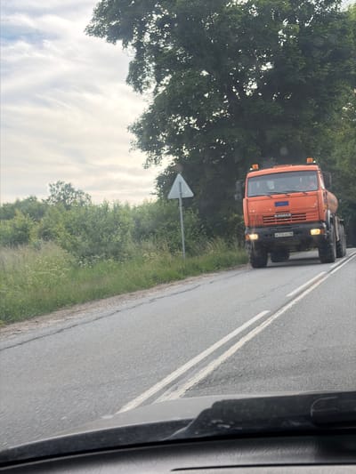 This media file captures an orange KAMAZ truck driving on a paved road. The truck is the central object, positioned on the right side of the frame and angled away from the viewer. The scene appears to be rural, with lush green trees and vegetation lining the left side of the road. A cloudy sky is visible above the trees. The road itself is asphalt with white lane markings. The perspective suggests the photo was taken from inside another vehicle, likely a car, as indicated by the dark interior element on the right edge of the frame. The time of day appears to be daylight, possibly late afternoon or early evening, given the soft light and shadows. There are no people clearly visible in the truck. The weather seems overcast. The image is slightly tilted, making the truck appear to be driving uphill.

Visible text on the truck includes "KAMAZ" on the grille, and a license plate with Cyrillic characters and numbers: "В 376 ХС 96". There are also some numbers "60" and "1282" on the front of the truck, possibly indicating a model or identification number. A white triangular sign on a pole is visible on the left side of the road, which could be a road sign, although its details are unclear. The setting of Kumachevo, Russia, provides the geographical context. The overall mood is neutral, documenting a common road scene.