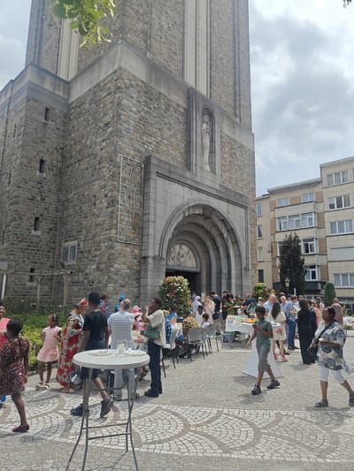 A group of people are gathered outside a stone church in Jette, Belgium, during what appears to be a community event or celebration. The church has a tall, imposing stone facade with an arched entrance adorned with sculptures. In front of the church, several tables are set up, some laden with food and drinks, suggesting a reception or outdoor meal. People of various ages and ethnicities are interacting, some standing and talking, others seated, and some are children walking or playing. Many are dressed in colorful attire. The ground is paved with a cobblestone pattern featuring decorative circular designs. The weather appears to be partly cloudy. Buildings are visible in the background, indicating an urban setting.