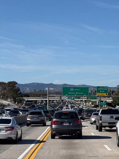 A congested multi-lane highway stretches into the distance under a clear blue sky, dotted with wispy clouds. Cars are densely packed, indicating heavy traffic. A green highway sign prominently displays "405 North Santa Monica," suggesting the location and direction of the freeway. Another sign points to "Exit 50B Jefferson Blvd." Mountainous terrain is visible in the background.