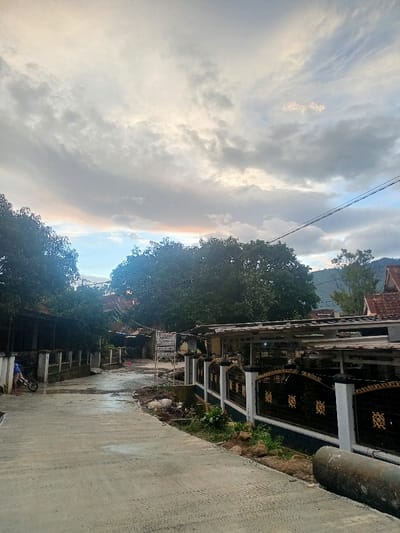 The image captures a tranquil street scene in Kecamatan Pangalengan, Indonesia, under an overcast sky. The time of day appears to be either late afternoon or early morning, hinted by soft, warm orange and pink hues visible on the horizon through breaks in the dense, grey clouds. The concrete road, which shows signs of being damp or recently wet, curves gently to the left, leading into the distance.
On the right side of the road, a property is bordered by a decorative black metal fence with golden scrollwork, featuring white pillars. Behind the fence, parts of a building or a shaded structure with a light-colored roof are visible, and further back, the traditional red-tiled roof of another house can be seen. Utility poles and power lines stretch across the upper right portion of the frame. Piles of dirt and small rocks are scattered along the base of the fence on this side, and a segment of a concrete pipe lies by the roadside.
To the left, a lush, dark green tree dominates the background, with other trees framing the left edge of the image. Partially obscured by a low wall on the left, a person is seen kneeling or bending next to a small blue motorcycle or scooter. A white informational sign stands prominently by the road near the center. The visible text on the sign indicates local government affiliation, reading: "PEMERINTAH KABUPATEN BANDUNG" (Government of Bandung Regency), "KECAMATAN PANGALENGAN" (Pangalengan Sub-district), followed by smaller text like "DESA..." (Village...) and "RT... RW..." (neighborhood administrative units). In the far background on the right, forested hills or mountains are discernible, suggesting a rural or mountainous setting. The overall atmosphere is quiet and subdued due to the muted lighting and heavy cloud cover.