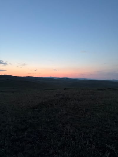 The image depicts a serene landscape at dusk, possibly in the vicinity of Kuseevo, Russia. The sky transitions from a pale blue at the top to hues of pink and orange near the horizon, indicating the setting of the sun. Soft clouds are scattered across the upper portion of the sky. Below the sky, rolling hills stretch into the distance, covered in dark, silhouetted vegetation. The foreground is dominated by a field of dry grass and low-lying plants, appearing dark and somewhat indistinct due to the low light conditions. There are no visible people, structures, or specific man-made objects in the scene, emphasizing the natural and rural character of the environment.