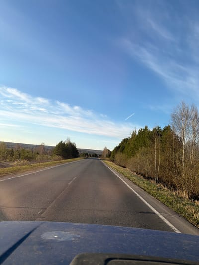 A slightly tilted shot depicts an asphalt road stretching into the distance, flanked by trees on the right and a blue sky with wispy clouds on the left. The lower right corner shows a portion of a blue vehicle, possibly the car from which the photo was taken. The location cannot be definitively determined as Балаки, Russia, from the visual information provided.