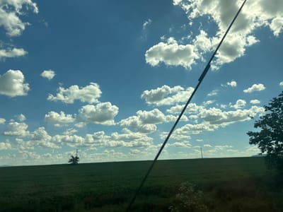 The image depicts a vast, green field under a bright blue sky filled with scattered white clouds. In the distance, a lone tree stands near a power pylon. A thin, dark pole, likely a fence post or utility pole, runs diagonally across the right side of the frame. The field appears to be cultivated with low-lying crops. The scene is characteristic of rural landscapes. The location of Lovets, Bulgaria is not explicitly identifiable from the visual elements in the image, as it is a general rural scene.