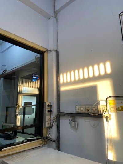 The image depicts an indoor scene with natural light casting distinct shadows on a white wall. The light appears to be entering from a window or opening, creating a pattern of parallel, elongated rectangles of light across the wall. In the foreground, a desk or counter is partially visible, with papers on its surface.
To the left, a glass partition or window reveals a glimpse into another room. Inside this room, a computer monitor is visible on a desk, along with some reflective surfaces. The lighting inside this adjacent room is different, with a cooler, blueish tone.
On the wall to the right, an electrical outlet strip with several sockets is mounted. Various plugs are inserted into these sockets, and numerous electrical cords are connected, some running along the wall and down towards the floor. Above the outlet strip, there are some yellow warning labels, one of which reads "HIDUPKAN LAMPU BILA DIPERLUKAN" (Turn on the lights when needed) and another with a lightning bolt symbol and "BAHAYA ARUS LISTRIK TEGANGAN TINGGI" (Danger High Voltage Electric Current).
The environment appears to be an office or a service area, possibly a consultation room or reception area, given the signage visible through the glass partition that includes "RUANG KONSULTASI" (Consultation Room). The overall atmosphere is utilitarian and functional. The presence of the sunlight suggests it is daytime. The specific location within Margoyoso, Indonesia, cannot be determined from the visual information alone; this detail would require external context.
