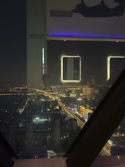 This media file captures a nighttime view of a cityscape from an elevated perspective, possibly a high-rise building, overlooking Jianye District in China. The image is oriented vertically.

The scene is dominated by the illuminated cityscape at night, with countless lights from buildings and streetlights creating a vibrant tapestry. A prominent element in the foreground is a dark, angled beam or frame, likely part of the window structure of the vantage point, which partially obscures the view. To the left, inside the viewing area, there is a wall with a dark silhouette of a person, appearing to be in a dynamic pose, and a vertical strip of blue light. There are also two rectangular, illuminated openings in the wall, one larger and one smaller.

The overall activity appears to be the observation of the city at night. The lighting suggests it is well after sunset. The weather is not discernible from the image. The combination of the city lights and the elevated viewpoint evokes a sense of urban scale and dynamism. There is no visible text in the media file.