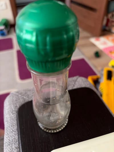 A close-up, slightly blurry, eye-level shot shows a clear glass spice grinder with a shiny green plastic lid, resting on a dark brown wooden surface. The grinder is partially filled with what appears to be fine brown spice particles. The background is out of focus, but shows a grey textured surface to the left, and hints of colorful items, possibly toys or floor mats, and a wooden surface to the right. There are no people visible in the image. The scene appears to be indoors, possibly in a residential setting. The image has a shallow depth of field, with the spice grinder being the primary subject in sharp focus. The location in the city of Mirnoye, Ukraine is not discernible from the image.