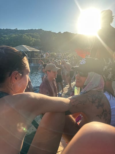 The image depicts an outdoor daytime gathering in Tisno, Croatia, characterized by strong sunlight and a large crowd. In the immediate foreground, two individuals are partially visible. On the left, the upper body of a person with dark hair, wearing sunglasses on top of the head and a large hoop earring, faces right. Their left arm is bent and prominent in the lower-middle frame. On the right, another individual, shirtless, faces left. This person wears sunglasses, a black baseball cap with a white "SS" logo resting on top of a light-colored, patterned headscarf, and has extensive dark tattoos on the left shoulder and upper back. Their right arm is bent and intersects with the first person's arm.

Behind these foreground figures, a body of water extends towards a wooden stage or DJ booth, which features a light-colored, possibly thatched roof and visible speakers. A large crowd of people is gathered around the stage, both on the rocky shoreline and standing in the shallow water. One individual in the mid-ground, wearing a light baseball cap and a reddish-brown covering, holds a blue can while standing near the water's edge. The scene is strongly backlit by a low sun on the right side of the frame, creating significant lens flare and silhouetting many background figures. Tree-covered hills rise behind the crowd and stage, under a clear blue sky.