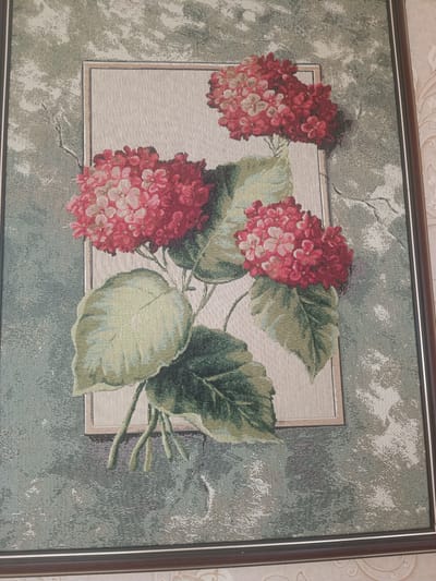 This is a close-up, eye-level, indoor shot of a framed tapestry. The tapestry depicts a cluster of hydrangeas with large green leaves. The hydrangeas are a deep red with some lighter pink and white highlights. The leaves are various shades of green, with prominent veining. The tapestry has a textured appearance, with a woven effect visible. The frame is a dark brown, and the tapestry is hanging on a wall with textured wallpaper. The lighting is soft and even, suggesting an indoor environment. There is no indication of the city of Mezhgorye, Russia, in the image.
