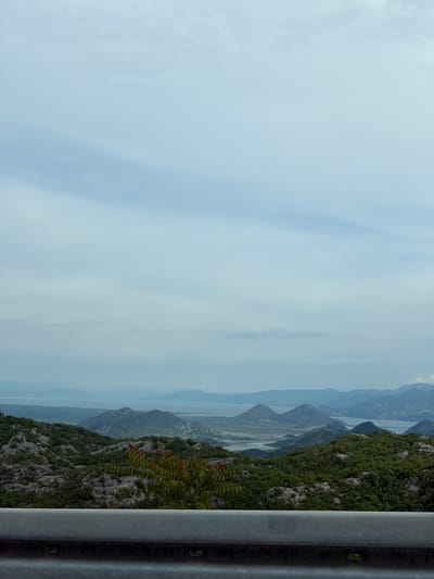 The image captures a vista from what appears to be a roadside in the vicinity of Gornja Brova, Montenegro. To the right, a gray metal guardrail runs vertically, its texture and details visible. Beyond the rail, the landscape slopes down, covered in lush green vegetation with rocky outcrops. In the distance, a series of mountains stretch out, shrouded in a light haze. Water bodies snake between the mountains, reflecting the sky. The sky itself is overcast, with soft blue hues peeking through the clouds.