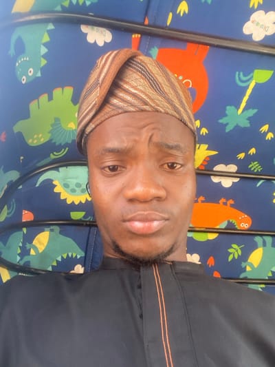 A young adult male, appearing to be of African descent, is depicted in a close-up portrait, presumably in Lapai, Nigeria. He has a light brown complexion, dark eyes, and a slight mustache and goatee. He wears a brown, striped traditional cap and a black garment, possibly a tunic, featuring a vertical orange stripe detail on the front. His expression is neutral with a slightly furrowed brow. The background consists of a blue fabric patterned with various cartoon dinosaurs (green, orange) and trees, interspersed with white clouds containing text such as "Dinooo!". Black metal bars are visible horizontally across the frame, suggesting the interior of a vehicle, possibly an auto-rickshaw.