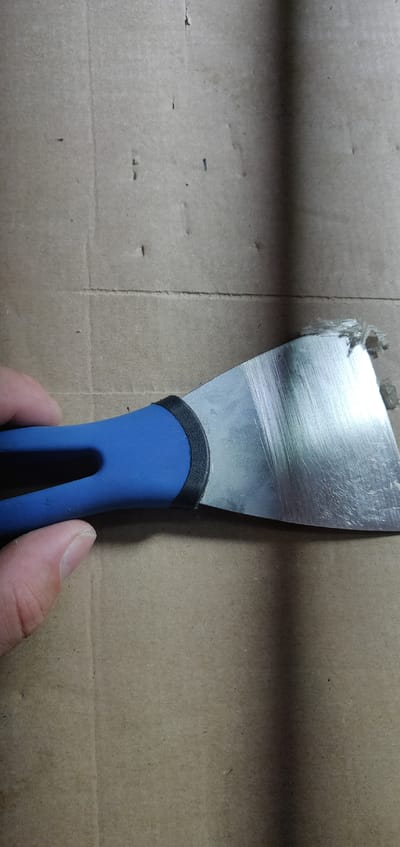 The image shows a close-up, overhead view of a blue and black handled putty knife held by a person's left hand against a textured brown cardboard surface. The putty knife's metal blade is angled upwards and to the right, with some fibrous material stuck to its leading edge. The cardboard has visible indentations and a vertical shadow cast from an unseen light source. The scene is indoors, and the location is not specified in the city of Lazear, United States.