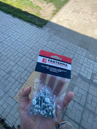A person's hand holds a clear plastic bag filled with metal screws. The bag is partially obscuring a red and black cardboard packaging with the brand name "FASTERRA" and Russian text. The packaging also contains contact information, including an address in Volgograd, Russia. The background consists of a gray, patterned brick walkway and a strip of green grass. The scene appears to be outdoors, possibly on a street or sidewalk. There is a shadow cast across the walkway, indicating sunlight. The image was captured in the city of Znamensk, Russia.