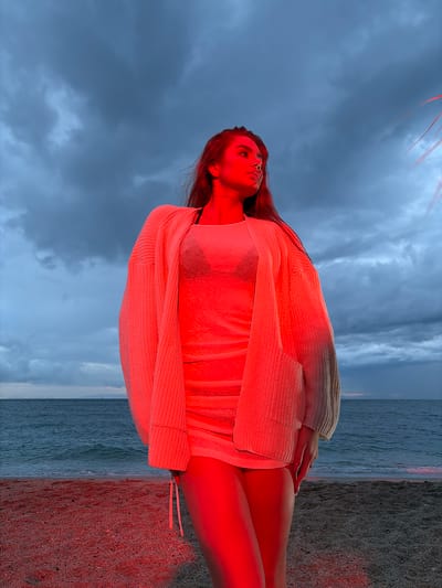 A young woman lies on a sandy beach in Alimos, Greece, with the Aegean Sea visible behind her. The sky above is overcast with blue-gray clouds. The woman is wearing a light-colored swimsuit covered by a cardigan. Her skin is illuminated with a red tint. The beach sand is also highlighted in red. The image appears to be taken from a low angle, looking up at the subject.