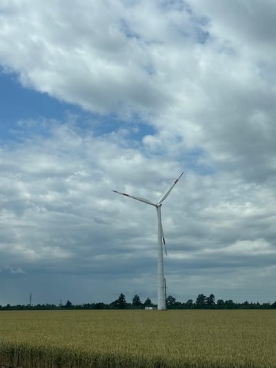 In Zarevo, Russia, a singular white wind turbine stands tall in a vast, golden field of wheat. The turbine's three blades are angled upwards, with red accents on their tips. The sky above is a dramatic canvas of grey and white clouds, interspersed with patches of bright blue. A line of dark green trees forms the horizon behind the field, with a power line tower visible in the distance. The overall scene conveys a sense of rural tranquility and renewable energy.