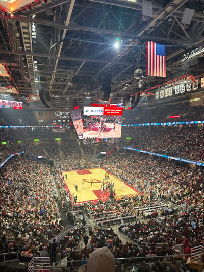 The image captures an overhead view of a basketball game taking place at Rocket Mortgage FieldHouse in Cleveland, Ohio. An American flag hangs prominently in the arena's rafters, next to the names and numbers of former players in a series of banners. A large video display board shows game highlights and advertisements. The basketball court is visible at the bottom of the frame, surrounded by densely packed stands filled with spectators.