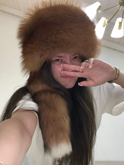 A single female subject is depicted in a close-up, front-facing indoor portrait, likely a selfie, captured in Vandœuvres, Switzerland. The subject, with long dark brown hair, is wearing a large, bushy brown fur hat, which appears to be an ushanka-style or similar winter cap, featuring a distinct, lighter-tipped tail-like extension draped over her right shoulder. She is also wearing a white t-shirt. Her right hand is positioned across the lower half of her face, with fingers pressed against or near her nose and mouth, implying a gesture of discomfort, playful disgust, or concealment. Visible accessories on her right hand include a ring with a white-colored stone on her index finger, a silver-toned ring on her middle finger, and a gold-colored, coiled bracelet on her wrist. Her fingernails are manicured with white tips. The background is predominantly a plain white wall and ceiling. In the upper right quadrant of the image, a portion of a modern chandelier with multiple light fixtures is visible, providing illumination to the scene.