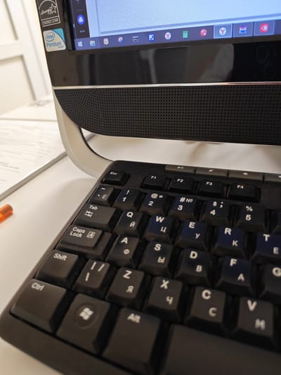 The image shows a close-up of a black keyboard in front of a computer monitor. The keyboard has both English and Cyrillic characters on its keys. Some keys visible include "Esc," "Tab," "Caps Lock," "Shift," "Ctrl," and several letter keys such as "W," "E," "R," "T," "S," "D," "F," "G," "Y," "U," "I," "O," "P," "A," "Z," "X," "C," and "V." There are also number keys "1" through "5." The monitor screen is partially visible, displaying icons. The background shows a white surface, possibly a desk, and a document. A small orange object, possibly a pen, is visible on the left side of the image. The overall scene depicts a standard computer workstation setup. There is no information or visual cues within the image to suggest the location is Krasnoselkup, Russia.