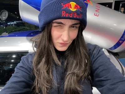 A woman with long brown hair wears a blue Red Bull beanie. She poses upside down, winking. Next to her is a large replica of a Red Bull can, displaying the energy drink's logo and branding. Behind her, partially visible, is a dark vehicle, possibly a car, with its windows reflecting the outside environment. The image has a casual, selfie-style feel.