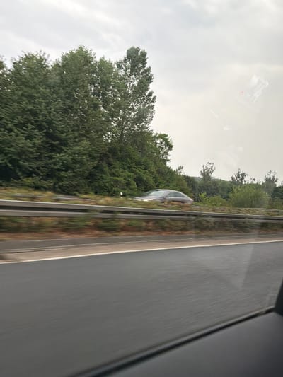 The image depicts a view from a vehicle moving on a highway in Tecklenburg, Germany. A dark-colored sedan is visible in motion on the adjacent lane, blurred due to the movement. The highway is flanked by a dense growth of green trees and vegetation. The sky is overcast and grey. The perspective suggests the photograph was taken from the passenger seat of a moving vehicle.